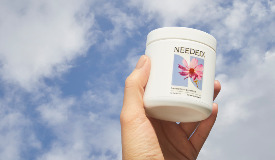 Hand holding Needed Prenatal Multi supplement jar against a blue sky